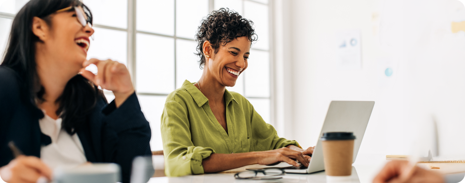 Duas mulheres sorrindo, sentadas na mesa de um escritório, usando notebook.